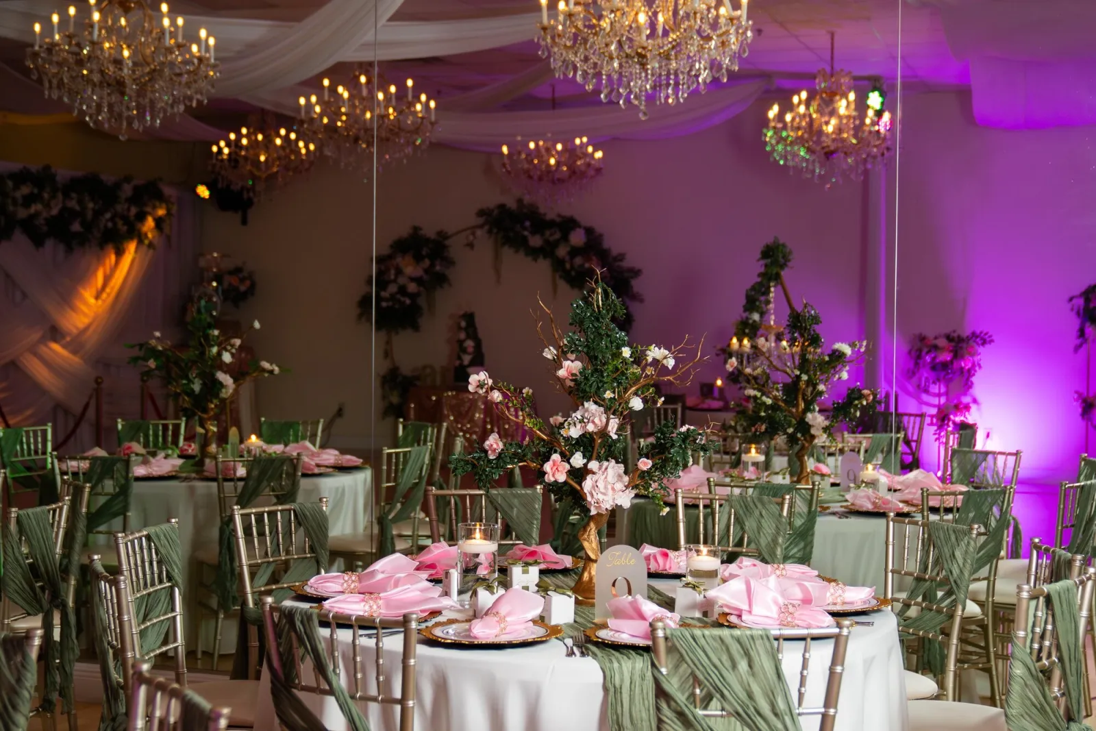 Full Nencini Ballroom pink and sage room design with chandeliers, floral centerpieces, gold chairs, candlelight, and purple uplighting